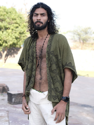 A man with curly hair and a beard stands outdoors wearing a green Cocoon Shrug, cream pants, a necklace, and a black smartwatch. Trees and a stone path can be seen in the background.