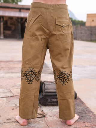 A barefoot person stands on stone, wearing sand yellow Tribal pants made from 100% cotton. The pants feature black hand blockprinted geometric patterns on the back and a right flap pocket. Outdoor walls and buildings are in the background.