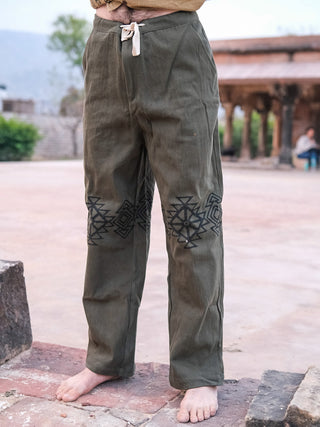 Wearing olive green Tribal pants made from 100% cotton, featuring black geometric embroidery on the thighs and a white drawstring, a person stands barefoot outdoors amid stone structures and lush greenery.