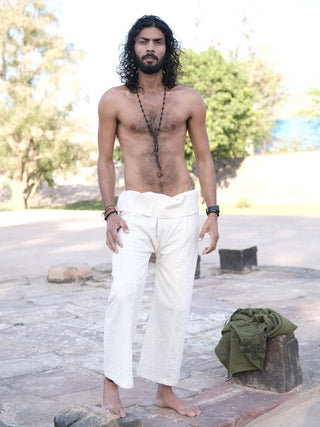 A barefoot man with long curly hair and a beard stands outside on stone, wearing white Ren Thai Pants and several necklaces, with a green cloth nearby. Trees and sunlight brighten the background.