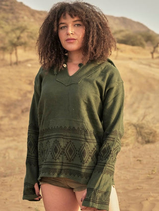 In a desert landscape, a curly-haired person wears a green Tribal Hoodie adorned with geometric patterns. The boho babe blends into the serene scene of sand, sparse trees, and a softly focused horizon.