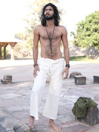 A barefoot, shirtless man with long curly hair and a beard stands on stone pavement outdoors, wearing Ren Thai Pants with a traditional silhouette, paired with beaded necklaces and bracelets. Trees and stone structures are behind him.