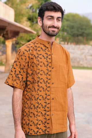 A man with short dark hair and a beard wears the Kunal Shirt, which features a bold leaf pattern on one side. Outdoors among trees and a stone wall, he smiles softly—showcasing the shirt as a statement piece.