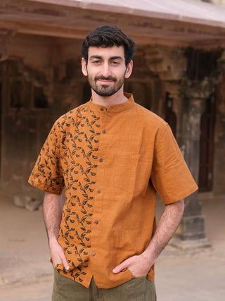A man with short dark hair and a beard stands outdoors, hands in pockets, wearing the Kunal Shirt—an orange statement shirt with a leafy pattern on one side—against an ancient stone building backdrop.