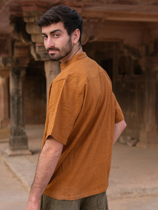 A man with short dark hair and a beard stands outdoors, turning to look back. He wears the orange-brown Kunal Shirt paired with green pants, set against old stone architecture—a true statement piece in a timeless setting.