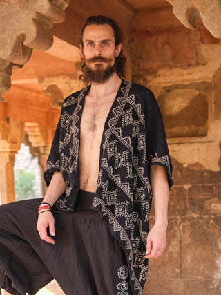 A bearded man with long hair stands in a stone archway, wearing the Cocoon Shrug—a hand block-printed black patterned open-front robe—paired with loose black cotton pants. The rustic background features weathered architecture.