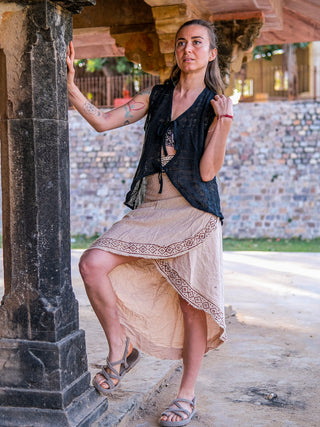 A tattooed woman outdoors rests her foot on a stone ledge, wearing sandals, a sleeveless black top, and the handmade block-printed Earthling Wrap Skirt. Sunlight highlights her face and the textured stone structure.