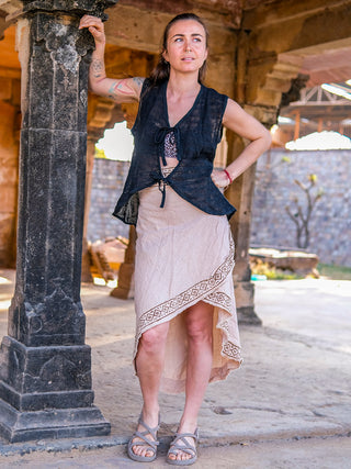 A woman with arm tattoos stands outdoors by a stone pillar, wearing a black sleeveless top and the Earthling Wrap Skirt—a beige Boho skirt featuring handmade block prints and a decorative border—looking thoughtfully into the distance.