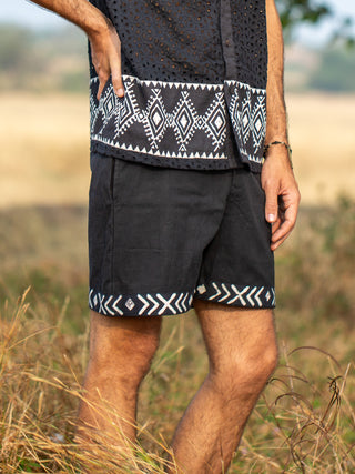 A person stands in tall grass wearing Tribal Shorts made from comfortable cotton, featuring white geometric patterns on the hems. Only their lower torso, legs, and one arm are visible.