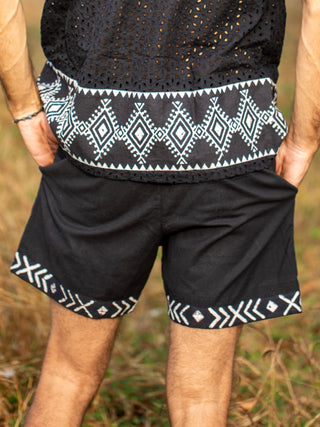 A person stands outdoors in black Tribal Shorts paired with a matching top, both edged with white geometric patterns. Made from comfortable cotton, the Tribal Shorts blend style and ease against a softly blurred grassy background.