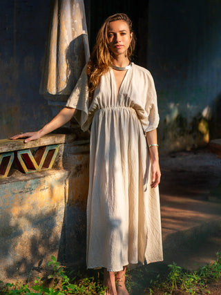 A woman wearing the Rusalka Maxi Dress in Natural stands barefoot outdoors, sunlight on her, one hand resting on a textured stone structure with greenery at her feet.