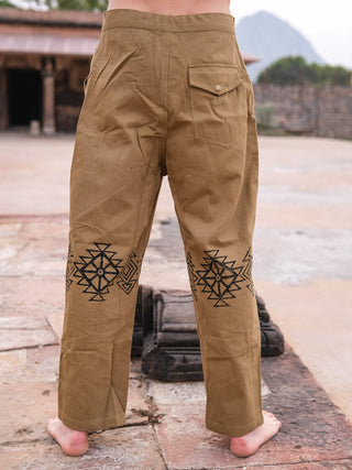 Barefoot outdoors, a person faces away wearing Tribal pants—100% cotton with black geometric hand-block prints on the back of the legs and a buttoned back pocket. Stone structures and distant mountains can be seen behind.