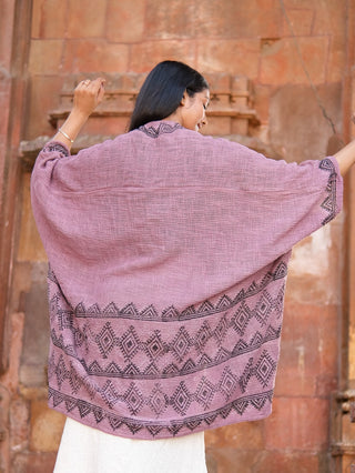 A woman faces away from the camera, arms raised, wearing the Cocoon Shrug—loose mauve cotton with black geometric hand block prints—set against a rustic stone wall.