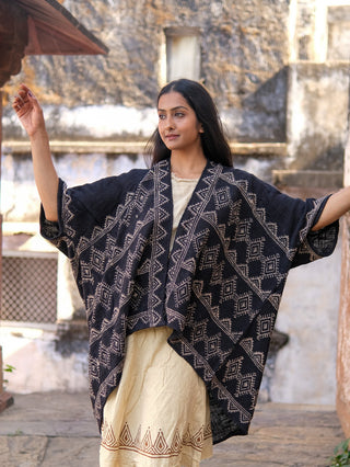 A woman stands outdoors in a hand block-printed, dark blue Cocoon Shrug over a cream dress with geometric designs, arms raised, with a rustic stone building behind her.