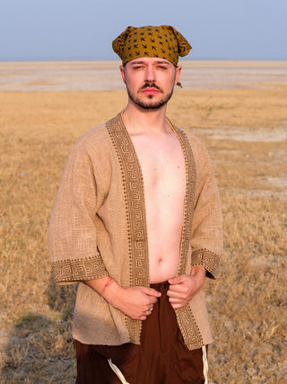 A man wearing the Atri Kimono, styled as a beige boho-inspired open-front cardigan, paired with brown pants and a patterned bandana, stands confidently in a dry, grassy field under clear skies.
