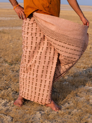 A person stands barefoot on dry grass, wearing the Rama Dhoti—a patterned beige and brown traditional garment—holding the cloth with one hand.