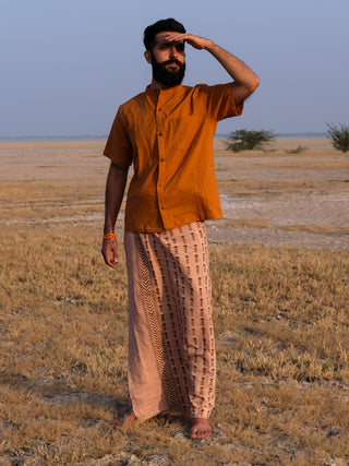 A bearded man in an orange shirt and a Rama Dhoti stands barefoot on dry grass, shading his eyes as he looks into the distance. The flat landscape stretches beneath a clear sky.