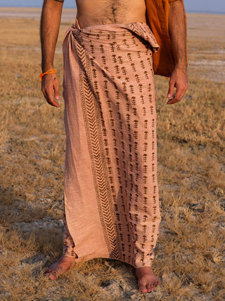 A person stands barefoot on dry grass, wearing the Rama Dhoti—a traditional peach garment with brown block print patterns—draped at the waist and revealing part of their upper body.