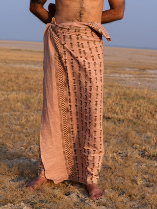 A person stands on dry grass wearing a light brown Rama Dhoti with black block print patterns, tied at the waist; their upper body is bare and face not visible.