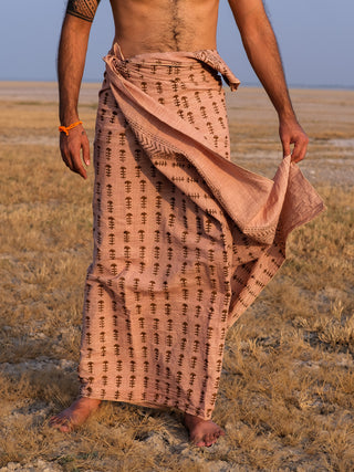A man stands barefoot on dry grass, wearing the Rama Dhoti—a patterned brown cotton traditional garment—wrapped at his waist. His upper body is bare, revealing a tattoo on his arm and an orange bracelet.