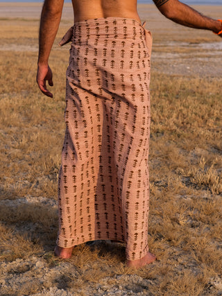 A person stands barefoot on dry grass, back to the camera, wearing a light brown Rama Dhoti block print wraparound garment tied at the waist.