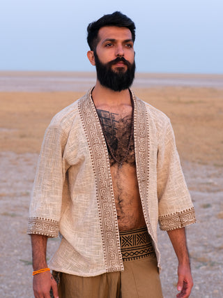 A man with a dark beard stands outdoors on a dry, open landscape. He wears the off-white, boho-inspired Atri Kimono open over his chest, showcasing a tattoo, paired with tan pants featuring a geometric waistband under a clear sky.