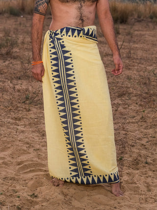 A person stands on sandy ground in the lemon yellow Jiva Dhoti, featuring a bold black geometric border. Only their torso and legs are shown, with a tattoo and orange bracelet, creating a subtly boho look.