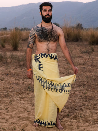 A bearded, tattooed man stands barefoot outdoors, holding one side of a Jiva Dhoti—traditional Indian attire with black boho patterns. Hills and dry grass form the backdrop.