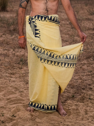 A person stands barefoot on sandy ground, wearing the Jiva Dhoti—a yellow traditional Indian garment with black geometric patterns. Their torso is bare, and one arm holds part of the wrap.