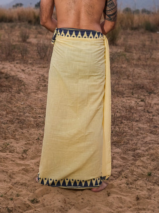 Outdoors on sandy ground, a person wears the Jiva Dhoti—a yellow traditional garment with a blue and yellow geometric border. Their back and upper arm tattoo are visible, set against dry grass and blurred trees.