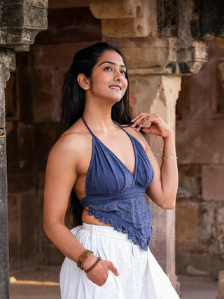 A woman stands outdoors by stone columns, wearing the stylish Titania Crop Top in blue with a white skirt. She smiles gently, looking to the side, with one hand in her hair and the other at her waist.