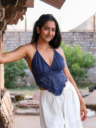 A woman with long dark hair smiles outdoors, leaning against a stone wall. She wears the Titania Crop Top in navy blue and loose white pants, surrounded by greenery and stone in the background.