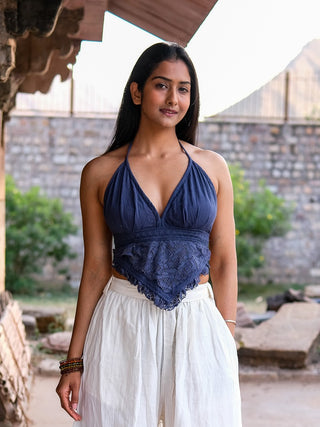 A woman with long dark hair stands outdoors in a blue Titania Crop Top and loose white pants, accessorized with bracelets. She looks relaxed against a stone wall and greenery backdrop.