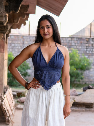 A woman with long dark hair stands outdoors in front of a stone structure, confidently posing with one hand on her hip. She wears the stylish Titania Crop Top in navy blue paired with white pants, surrounded by lush greenery.