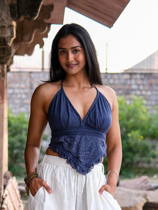 A smiling woman with long dark hair wears a Titania Crop Top and white skirt while standing outdoors by a stone structure, with greenery and a brick wall in the background.