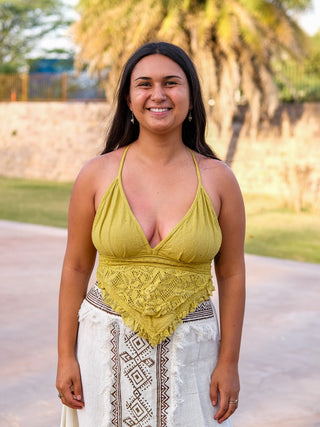 A woman with long dark hair smiles outdoors, wearing a yellow Titania Crop Top and a white geometric-patterned skirt. A palm tree and stone wall create a beautiful backdrop.