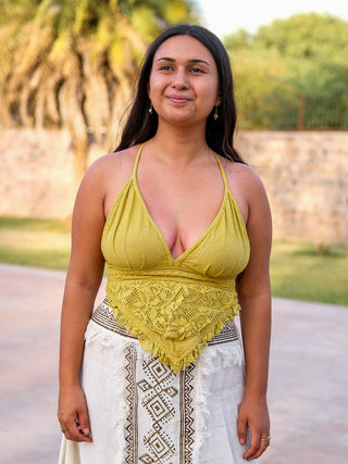 A woman with long dark hair stands outdoors, smiling in a lemon yellow Titania Crop Top with lace details and a white skirt with geometric patterns. Green trees and a stone wall complete the scene.