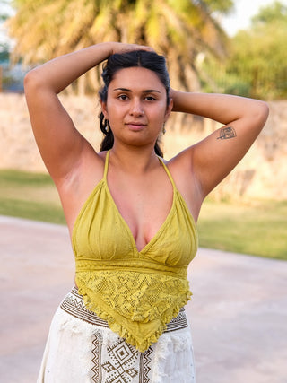 A person stands outdoors in a Titania Crop Top with arms raised behind their head, confidently facing the camera. Lush greenery and a palm tree form the background.