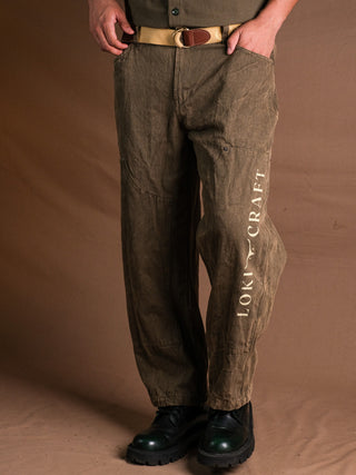 Someone stands against a brown background, shown from mid-torso down, wearing Chopper Pants in olive green with vintage styling and HOLY CRAFT printed on the left leg, paired with a tan belt and black boots.