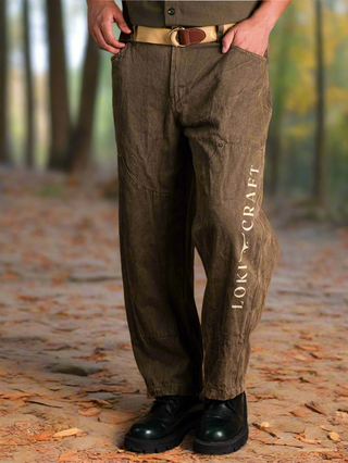 A person stands on a forest path wearing vintage-style Chopper Pants, featuring LOKY CRAFT branding, paired with a beige belt, brown shirt, and black boots. The blurred background shows trees and autumn leaves on the ground.