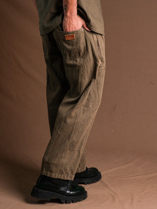 A person wears olive green Chopper Pants featuring a vintage-style back pocket label and black chunky-soled shoes, standing on a brown fabric floor with a matching brown backdrop.