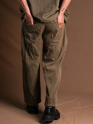 A person faces away from the camera, hands in the back pockets of loose olive green Chopper Pants with a subtle stonewash, matched with a green shirt and black boots against a plain brown backdrop.