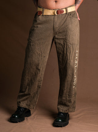 A person models olive-green Chopper Pants with visible stitching, vintage stonewash, and LOOK CRAFT printed vertically on one leg, styled with a beige belt and black chunky shoes against a brown background.