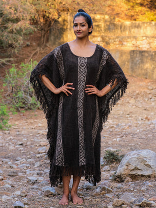 Standing confidently on rocky terrain, a person showcases their boho style with the Vama poncho, crafted from premium knitted fabric. The dark, fringed design with vertical stripes complements their short hair tied back as they rest their hands on hips amidst the dry ground and sparse vegetation.