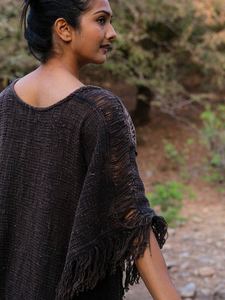 A woman outdoor stands in a dark, fringed Vama poncho with textured design. Her hair is tied back as she gazes sideways, highlighting the boho style amidst trees and rocky terrain.