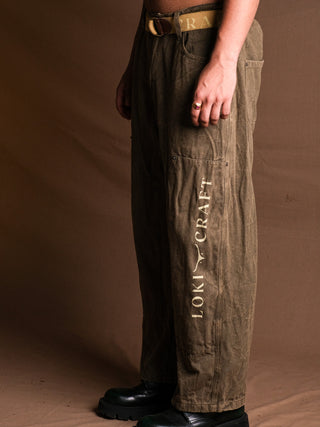 Against a brown background, a person showcases the Chopper Pants in olive green with LOKI CRAFT printed vertically on the leg, styled with a brown belt and black shoes for a subtle vintage look. Only the lower half of their body is visible.
