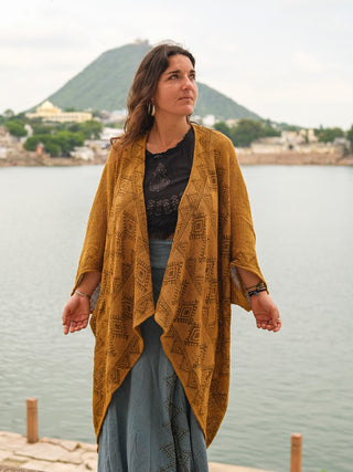 A woman wearing the ginger Cocoon Shrug stands outdoors by a lake with hills and buildings in the background, gazing to the side on a cloudy day.