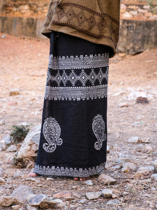 A person stands on rocky ground wearing a Vasuki dhoti, a long black skirt with white intricate and paisley patterns, reminiscent of Rajasthani handblock print, paired with a brown patterned cloth draped over their shoulders.