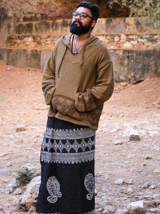 A man with a beard, glasses, and dark hair stands on rocky ground outdoors. Hes wearing a brown hooded sweater paired with the Vasuki dhoti. The background showcases dry vegetation and a stone wall.