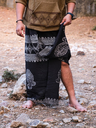 A person stands barefoot on rocky ground, partially lifting the black Vasuki dhoti adorned with intricate white tribal patterns. They wear a Rajasthani handblock print garment and have a wristwatch on their left hand, against an earthy backdrop scattered with rocks.